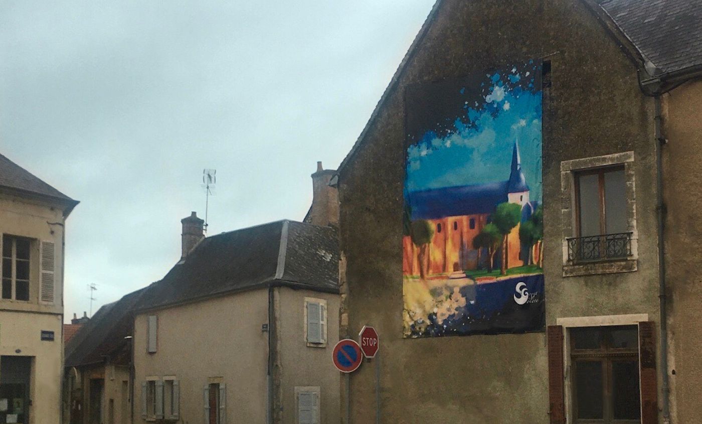photo de l'installation dans la ville de Charost d'une des peintures numérique représentant l’église de Charost, fresque réalisée par Solène Goget en 2022. Vue de trois quart.