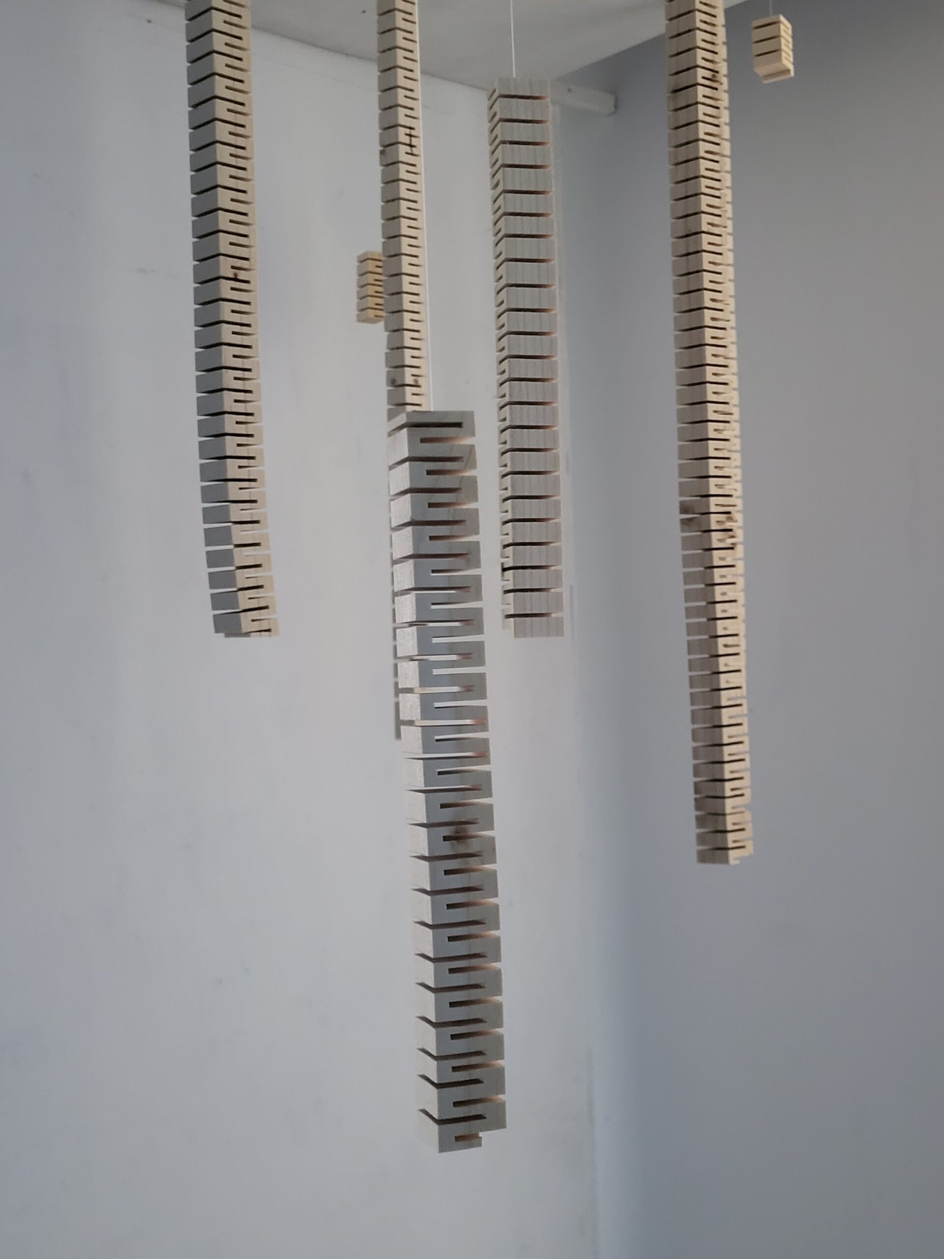 installation sonore immersive Suspension traversante de Solène Goget (2021-2022). Dans une salle blanche, plusieurs modules en bois articulé, suspendus au plafond par des fils fins, descendent verticalement à différentes hauteurs. La personne, placée au centre, touche l’un des modules, créant un lien direct avec la structure. L’œuvre, inspirée de la lutherie, diffuse des vibrations infrabasses de 85 Hz, invitant le spectateur à ressentir physiquement les ondes sonores dans son corps, transformant la traversée de l’espace en expérience méditative et sensorielle.