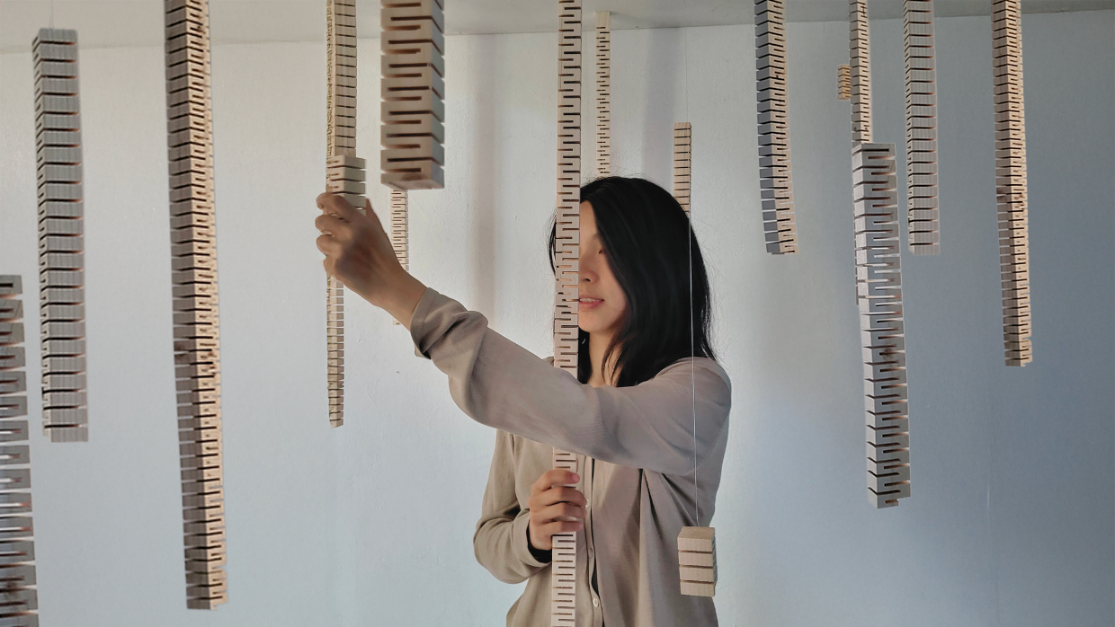 Une personne interagit avec l’installation sonore immersive Suspension traversante de Solène Goget (2021-2022). Dans une salle blanche, plusieurs modules en bois articulé, suspendus au plafond par des fils fins, descendent verticalement à différentes hauteurs. La personne, placée au centre, touche l’un des modules, créant un lien direct avec la structure. L’œuvre, inspirée de la lutherie, diffuse des vibrations infrabasses de 85 Hz, invitant le spectateur à ressentir physiquement les ondes sonores dans son corps, transformant la traversée de l’espace en expérience méditative et sensorielle.