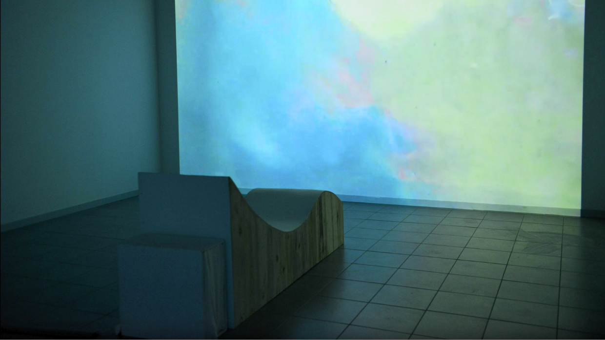 Installation Mélodie du corps de l’artiste Solène Goget. Une personne est allongée sur une structure en bois sinueuse, pensée comme une table vibratoire. Les bras ouverts, le corps épouse la forme sculptée de l’installation et devient caisse de résonance. Baigné dans une lumière bleue et face à une projection évoquant une surface aquatique, le spectateur vit une immersion sensorielle où le son se diffuse à travers la peau, le dos et la colonne vertébrale, invitant à un moment suspendu entre abandon et voyage intérieur.