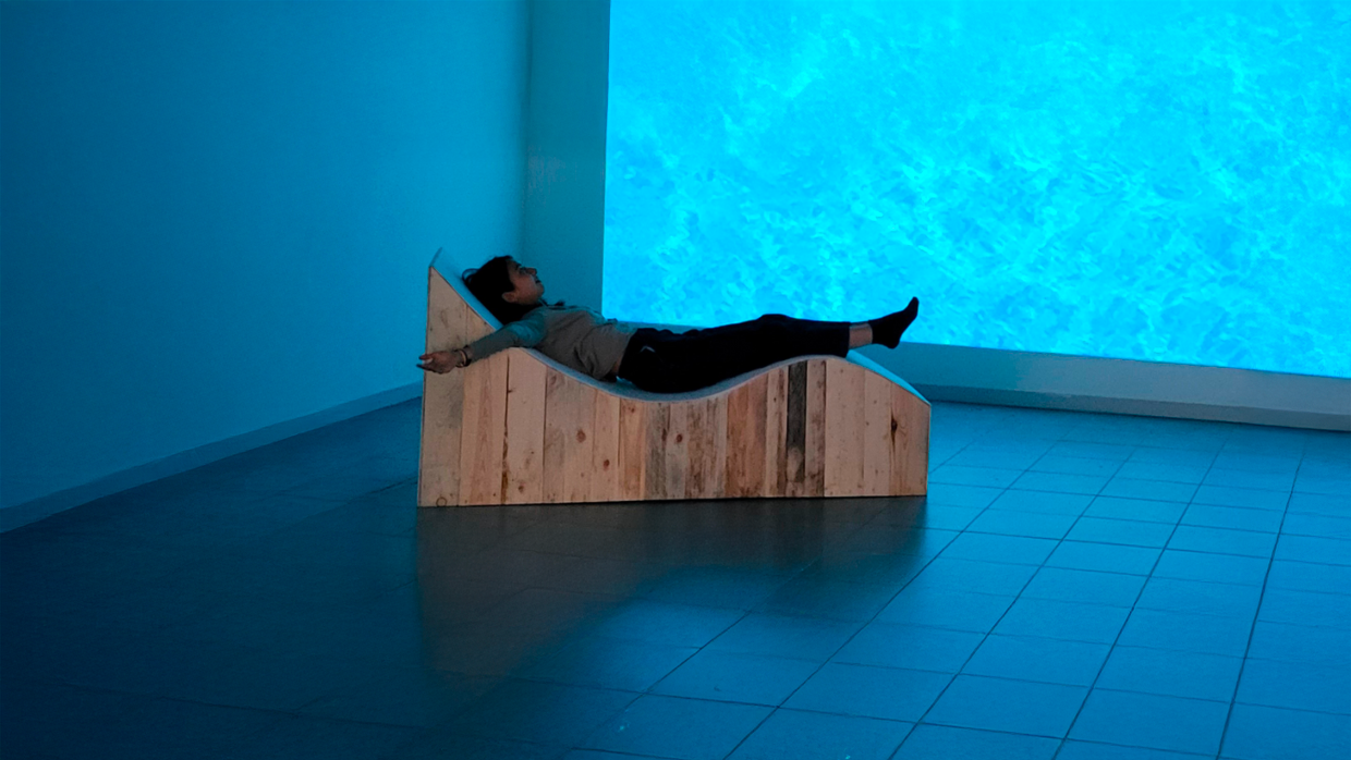 Installation Mélodie du corps de l’artiste Solène Goget. Une personne est allongée sur une structure en bois sinueuse, pensée comme une table vibratoire. Les bras ouverts, le corps épouse la forme sculptée de l’installation et devient caisse de résonance. Baigné dans une lumière bleue et face à une projection évoquant une surface aquatique, le spectateur vit une immersion sensorielle où le son se diffuse à travers la peau, le dos et la colonne vertébrale, invitant à un moment suspendu entre abandon et voyage intérieur.