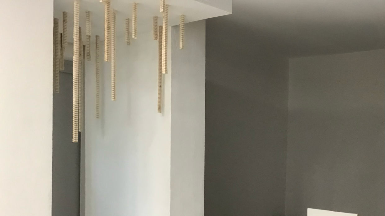 installation sonore immersive Suspension traversante de Solène Goget (2021-2022). Dans une salle blanche, plusieurs modules en bois articulé, suspendus au plafond par des fils fins, descendent verticalement à différentes hauteurs. La personne, placée au centre, touche l’un des modules, créant un lien direct avec la structure. L’œuvre, inspirée de la lutherie, diffuse des vibrations infrabasses de 85 Hz, invitant le spectateur à ressentir physiquement les ondes sonores dans son corps, transformant la traversée de l’espace en expérience méditative et sensorielle.