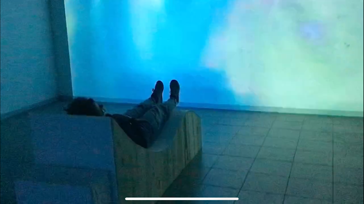 Installation Mélodie du corps de l’artiste Solène Goget. Une personne est allongée sur une structure en bois sinueuse, pensée comme une table vibratoire. Les bras ouverts, le corps épouse la forme sculptée de l’installation et devient caisse de résonance. Baigné dans une lumière bleue et face à une projection évoquant une surface aquatique, le spectateur vit une immersion sensorielle où le son se diffuse à travers la peau, le dos et la colonne vertébrale, invitant à un moment suspendu entre abandon et voyage intérieur.