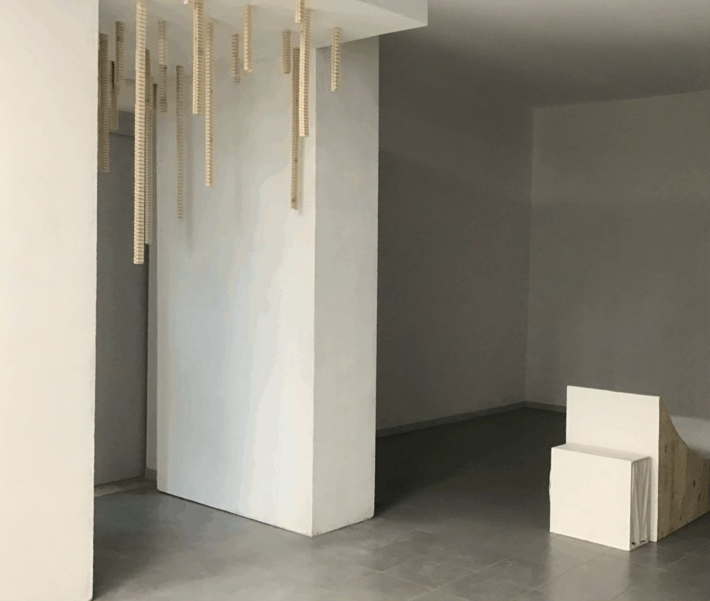 installation sonore immersive Suspension traversante de Solène Goget (2021-2022). Dans une salle blanche, plusieurs modules en bois articulé, suspendus au plafond par des fils fins, descendent verticalement à différentes hauteurs. La personne, placée au centre, touche l’un des modules, créant un lien direct avec la structure. L’œuvre, inspirée de la lutherie, diffuse des vibrations infrabasses de 85 Hz, invitant le spectateur à ressentir physiquement les ondes sonores dans son corps, transformant la traversée de l’espace en expérience méditative et sensorielle.